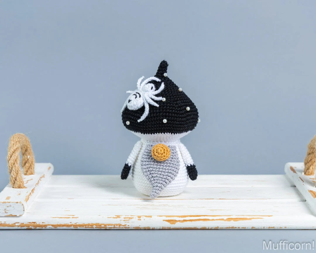 A crochetedmushroom gnome with a black and white spotted cap, white body, and a yellow-orange button, displayed on a rustic white wooden shelf against a light blue-gray background.