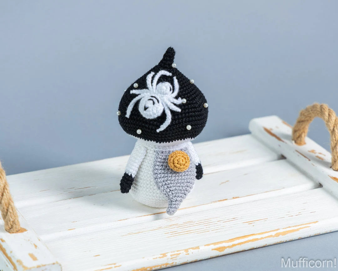 A crocheted amigurumi mushroom gnome with a white spider motif, standing on a white wooden platform against a soft blue-gray background.