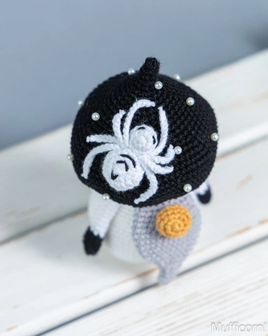 A handmade crocheted mushroom gnome with a black and white body, a white spider design on its head, and a small yellow nose, resting on a white wooden surface.