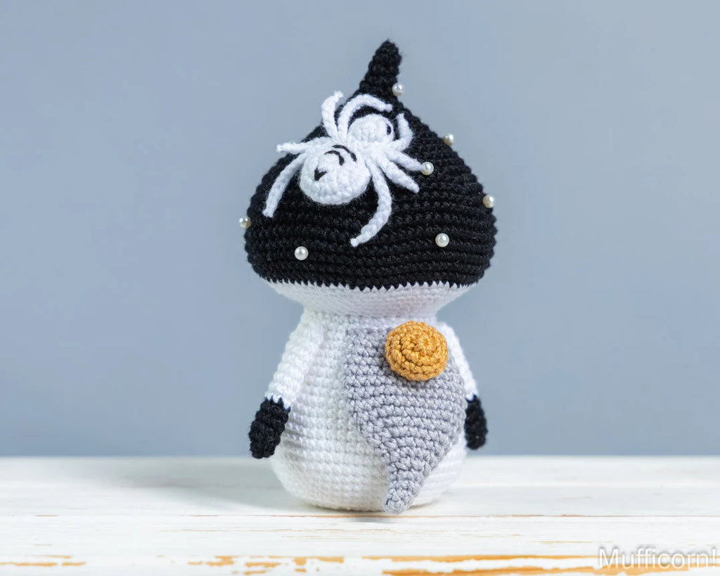 A handmade crocheted mushroom gnomer, featuring a black and white body with a white spider detail on its head and a yellow-orange circular accent on the chest, set against a soft gray background.