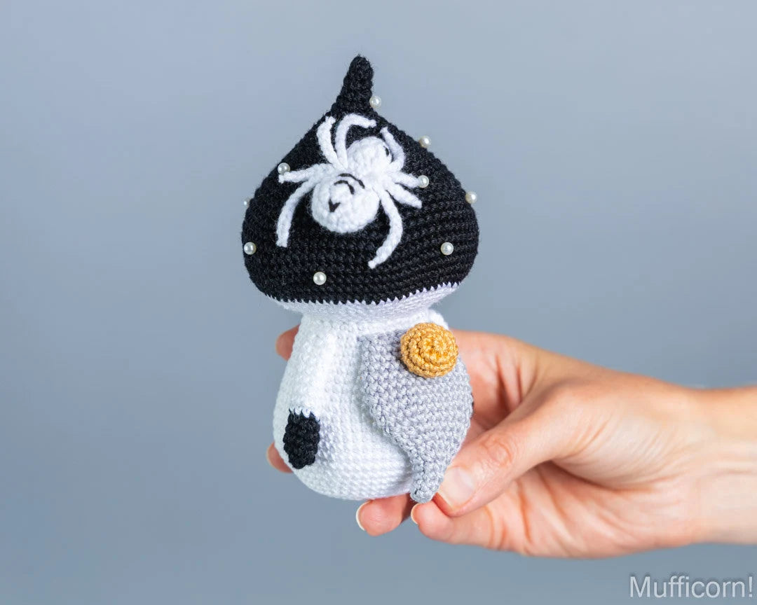 A handmade crocheted mushroom gnome with a black cap, white spider detail, and a white body, held in a person's hand against a solid gray background.