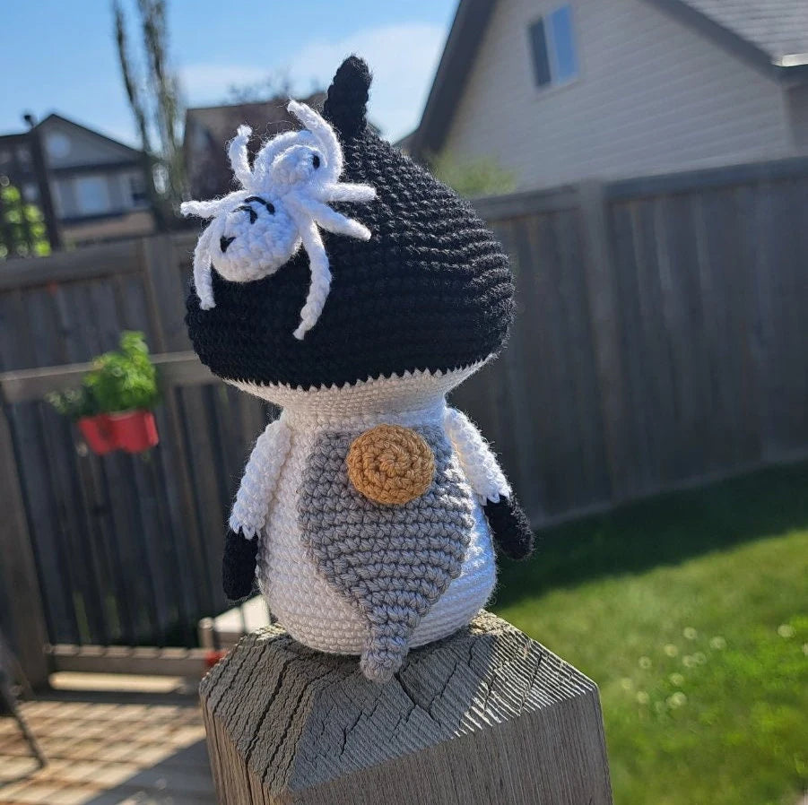 A crocheted mushroom gnome with a white spider on its forehead and a gray belly patch, sitting on a wooden post in a backyard.