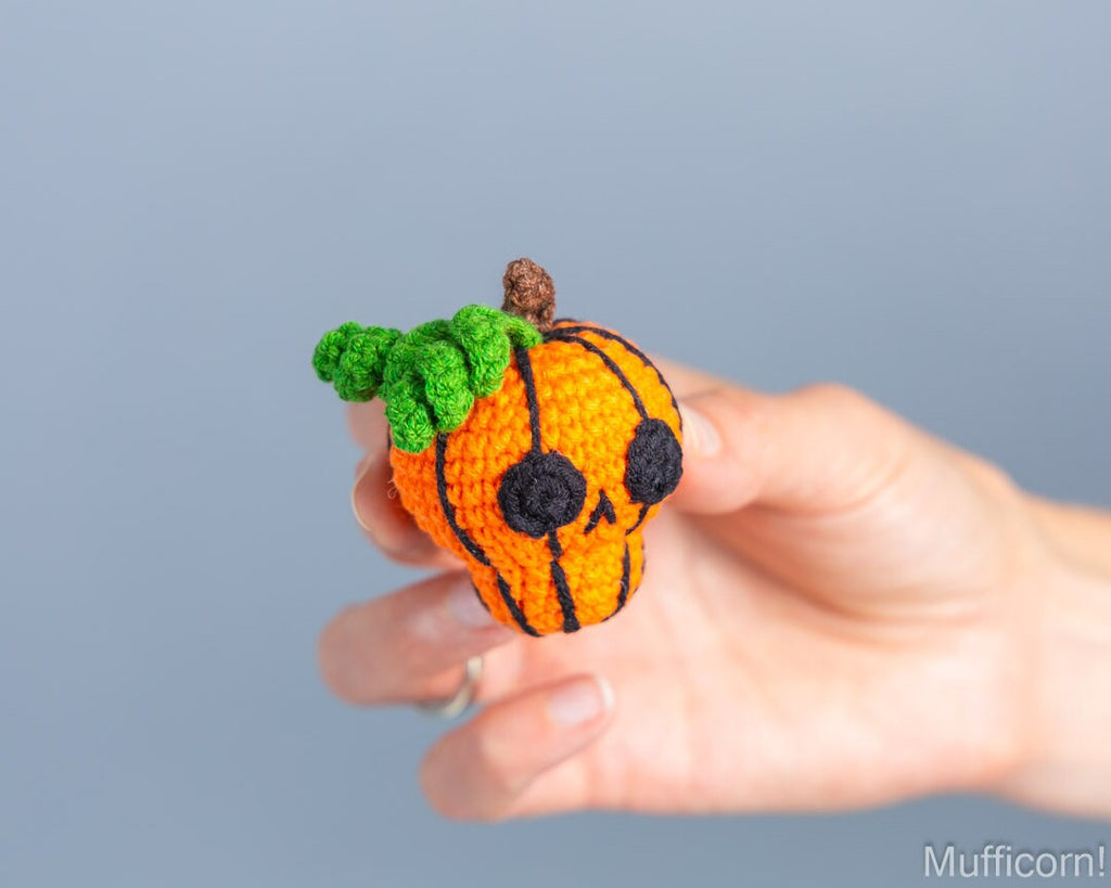 A hand holding a small crocheted pumpkin with a jack-o'-lantern face and green leafy top against a soft blue-gray background.