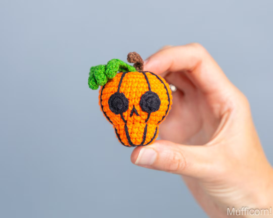 A hand holds a crocheted pumpkin-shaped skull with a green leafy top, black embroidered face details, and orange vertical stripes against a plain light gray background.