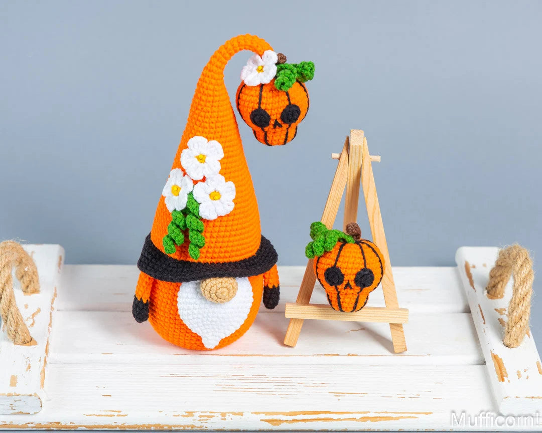 A crocheted orange gnome with a tall pointed hat decorated with white flowers and green leaves, standing beside a small wooden easel holding a matching crocheted skull pumpkin. The scene is styled on a distressed white wooden surface against a soft gray background.