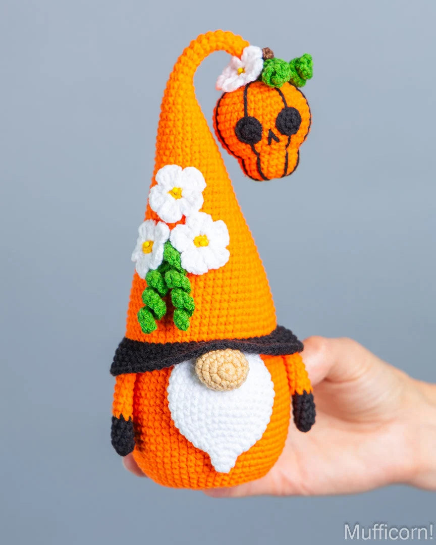 A crocheted orange gnome doll with a tall pointed hat decorated with white flowers and green leaves, a black band, a white beard, and a small hanging pumpkin, being held in a person's hand against a plain gray background.