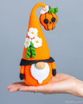 A crocheted orange gnome with a tall pointed hat, white flowers, green leaves, a black band, and a white beard, and orange skull being held in a person's hand against a plain gray background.