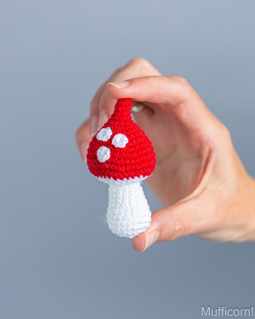 A hand holding a small crocheted mushroom toy with a red cap, white spots, and a white stem against a plain gray background.