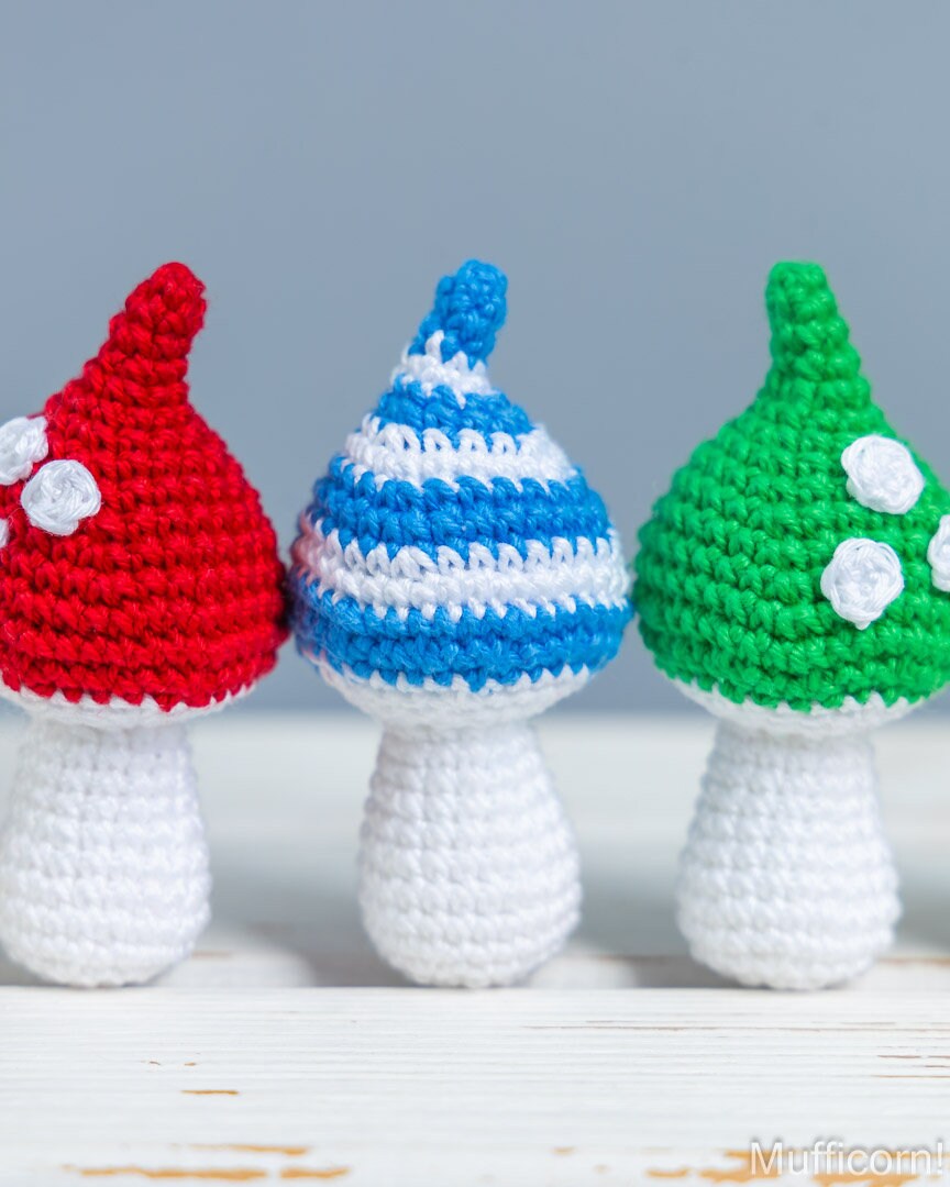Three crocheted mushroom figurines in red, blue-and-white, and green with white stems and spots, arranged on a white wooden surface against a soft gray background.