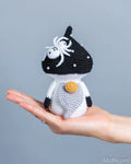 A small crocheted mushroom gnome with a black and white body, white spider detail on the head, and a gold-colored flower on the chest, resting on an open hand against a solid gray background.