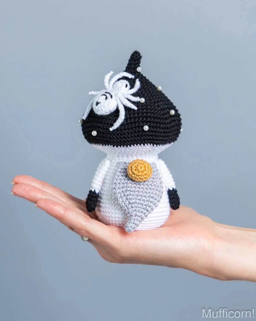 A small crocheted mushroom gnome with a black and white body, white spider detail on the head, and a gold-colored flower on the chest, resting on an open hand against a solid gray background.