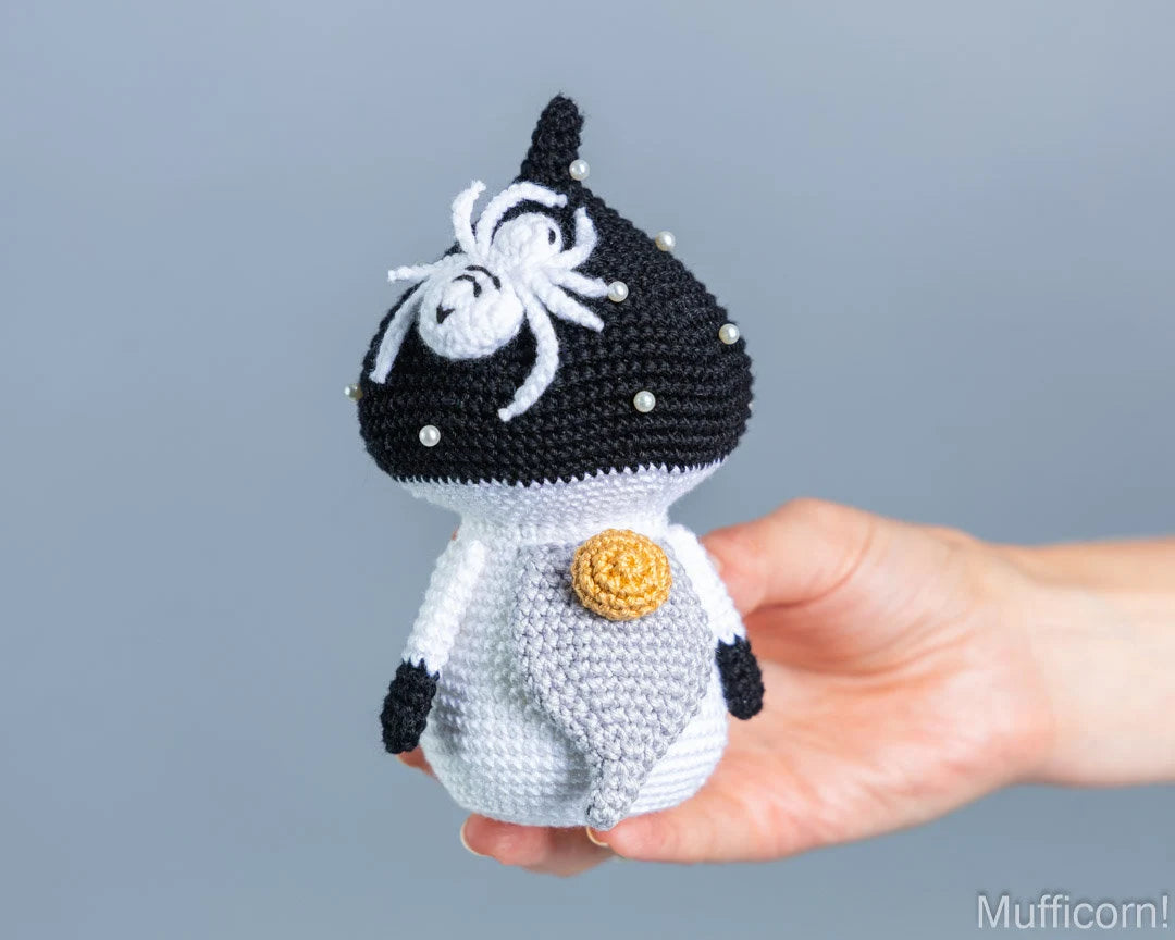 A hand holds a crocheted mushroom gnomet a plain light gray background. The toy is black and white with a yellow circular accent on its chest and small white bead details on its head.