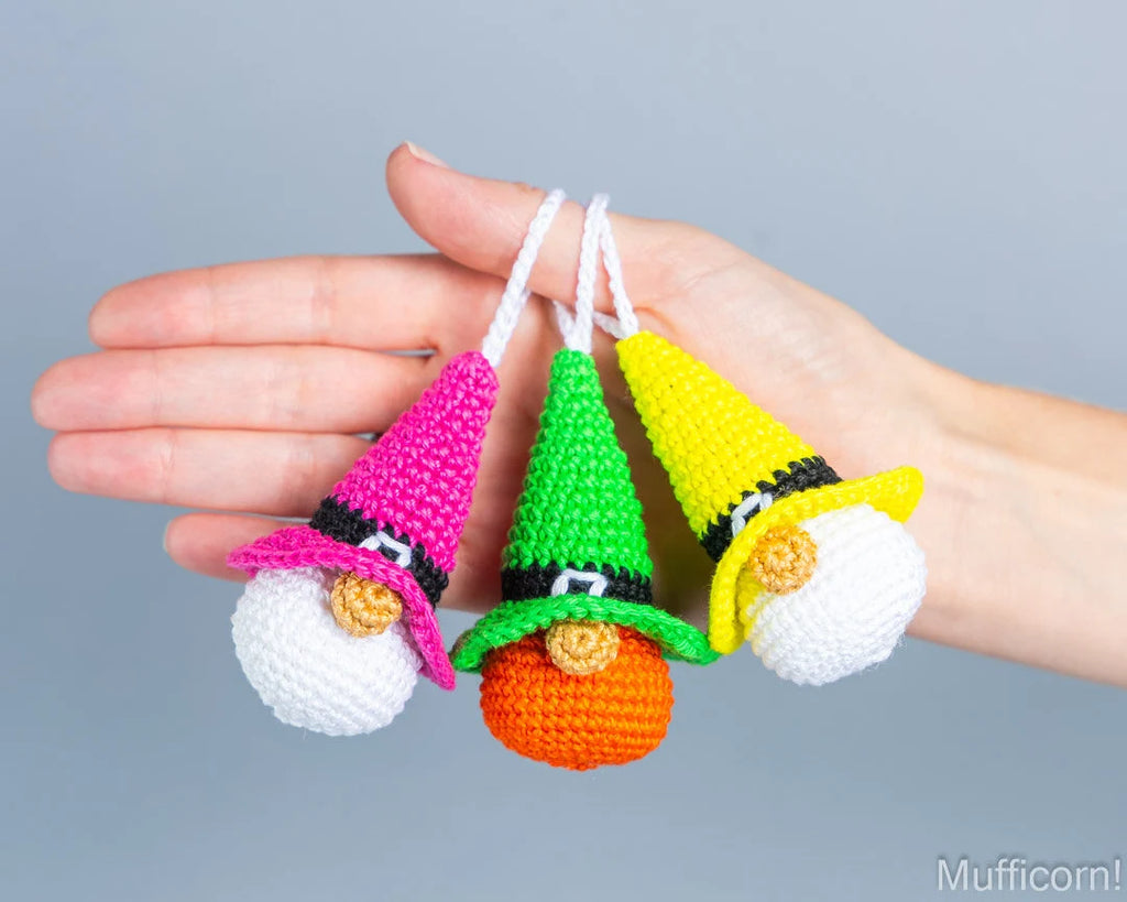 Three crocheted gnomes in witch hats ornaments held in a person's hand against a plain gray background. The ornaments have white beards, pointed hats in pink, green, and yellow, black bands, orange pots, and white hanging strings.