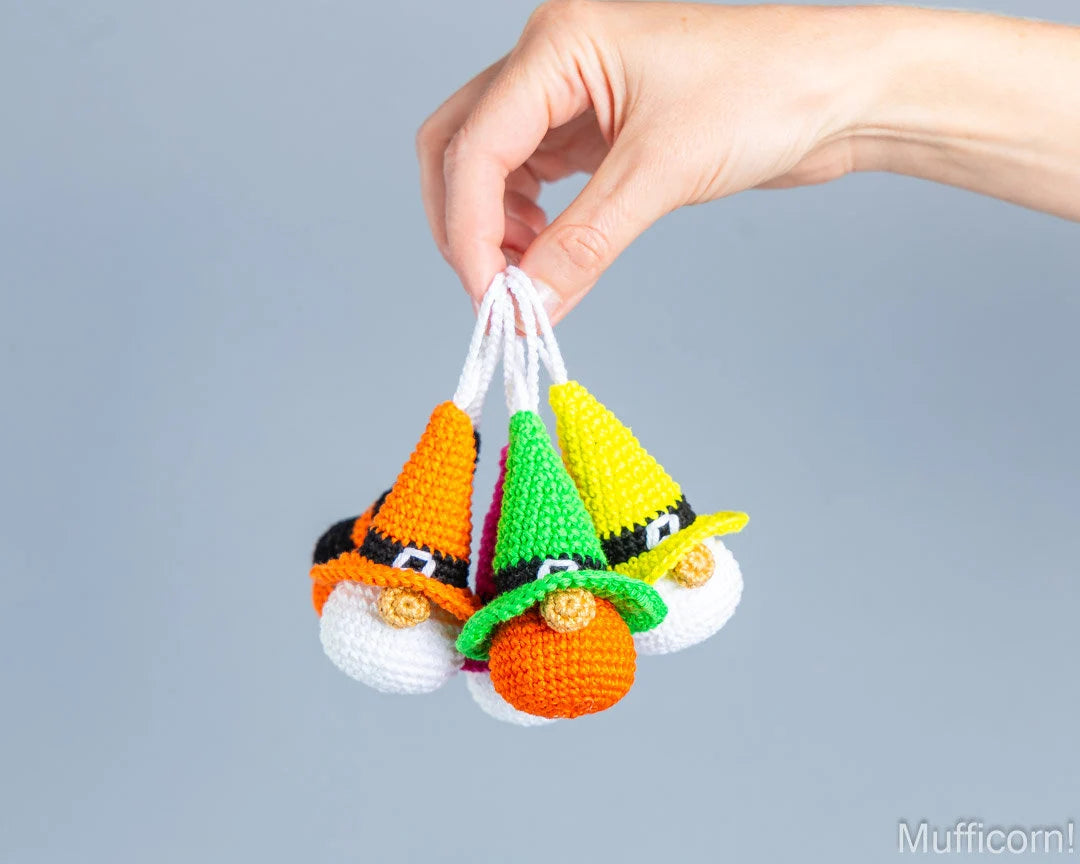 Five small crocheted gnome ornaments in orange, green, and yellow hats are held by a person's hand against a plain light gray background.