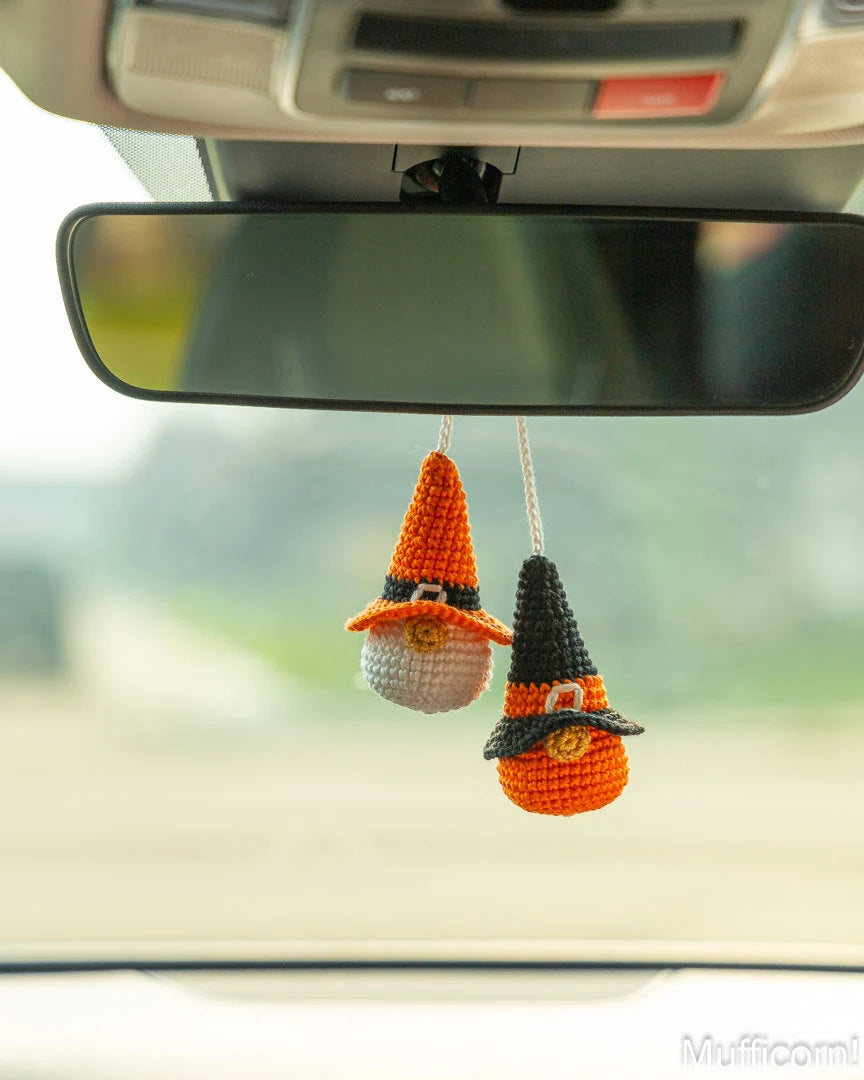 Two crocheted witch hat car air fresheners hanging from a car's rearview mirror. One is orange and white, and the other is orange, black, and white, with a blurred outdoor background visible through the windshield.