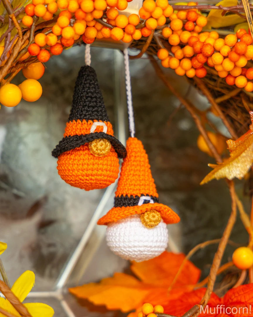 Two crocheted pumpkin gnome ornaments hanging from a fall wreath decorated with orange berries and autumn leaves. The ornaments feature orange and black pointed hats, black bandanas, and one has a white beard.