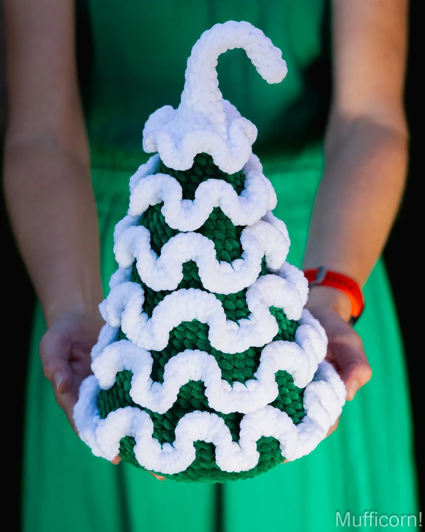 A person in a bright green dress holds a crocheted Christmas tree ornament. The ornament is dark green with white wavy yarn trim and a white curly top.