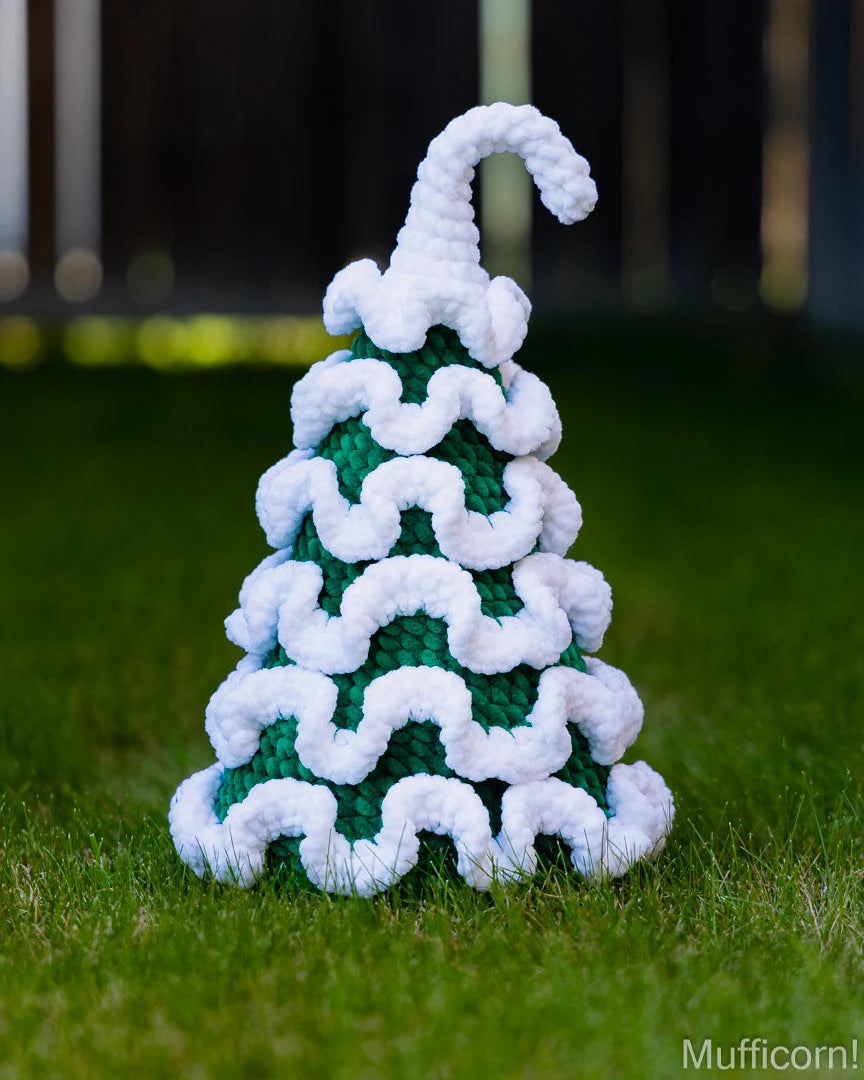 A crocheted Christmas tree ornament with a green body and white wavy trim, standing on grass outdoors with a blurred fence in the background.