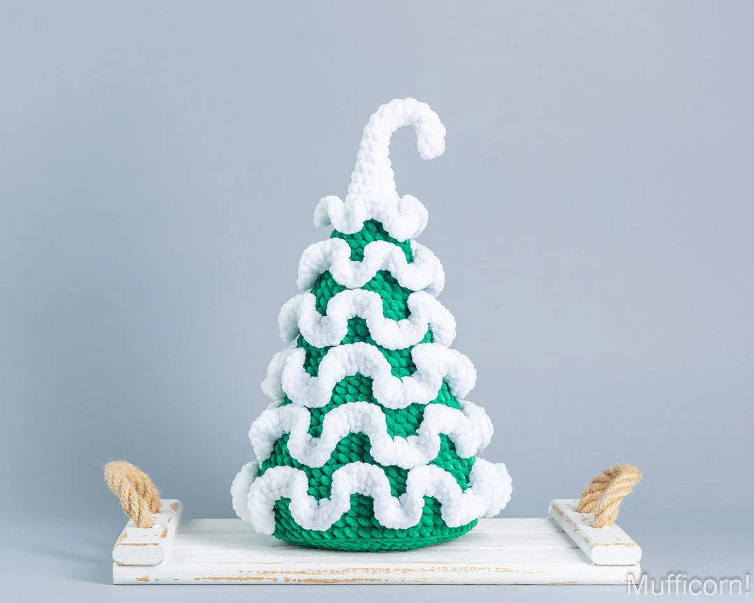 A crocheted Christmas tree ornament in green and white, styled on a white wooden tray with rope handles against a solid gray background.