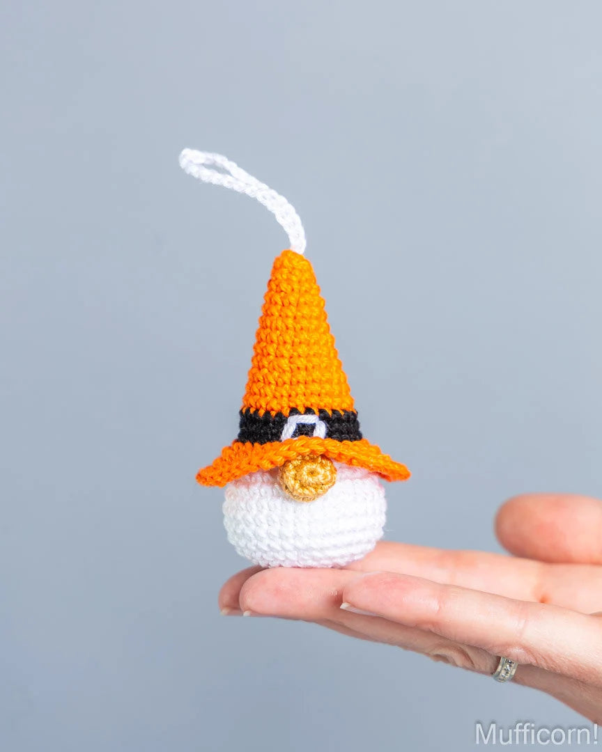 A small crocheted witch hat ornament being held on a person's fingers against a plain light gray background. The ornament has an orange pointed hat, a black band, a white face area, a gold circular nose detail, and a white hanging loop.