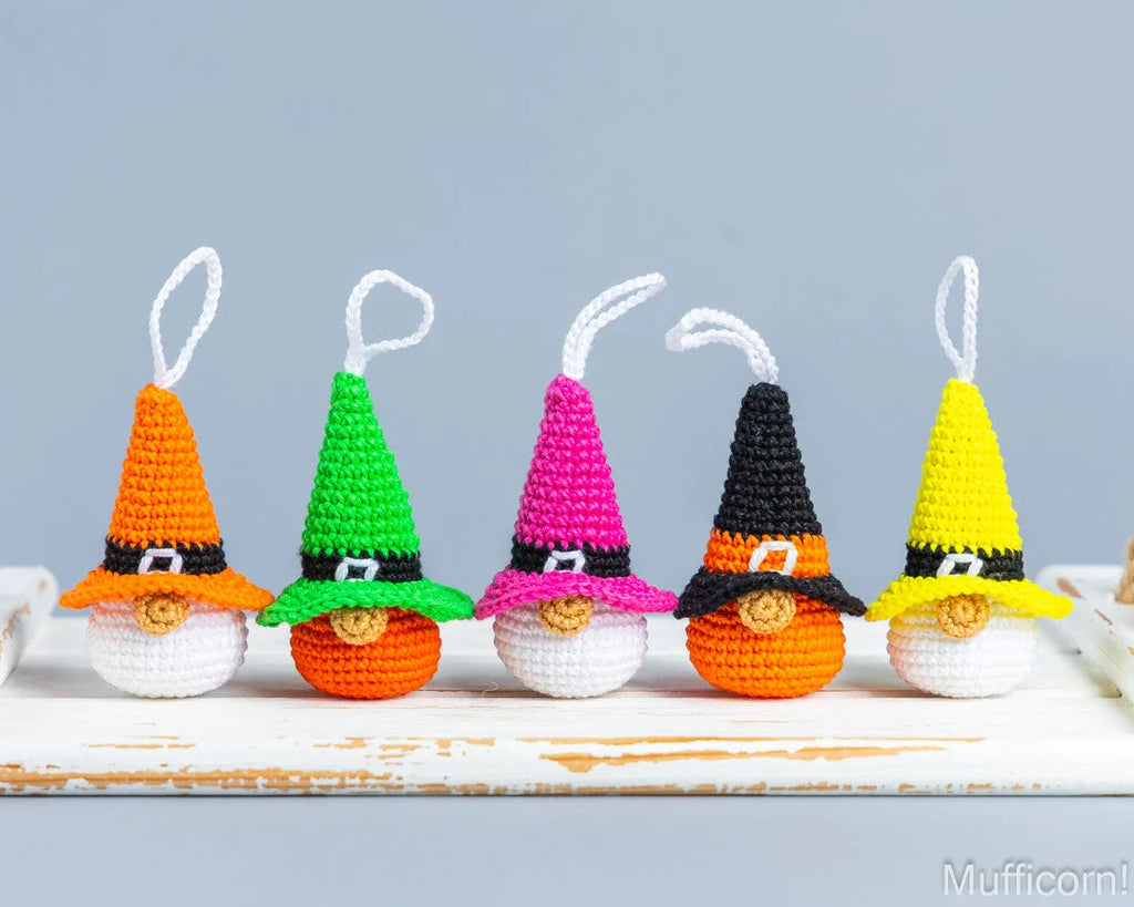 Five small crocheted Halloween gnome ornaments in different hat colors (orange, green, pink, black, and yellow) lined up on a white rustic wooden surface against a light blue-gray background.