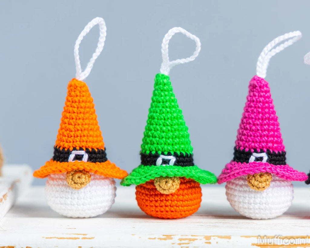 Three crocheted gnome ornaments wearing pointed hats in orange, green, and pink, with black bands, white beards, and white hanging loops, displayed on a white surface against a light gray background.