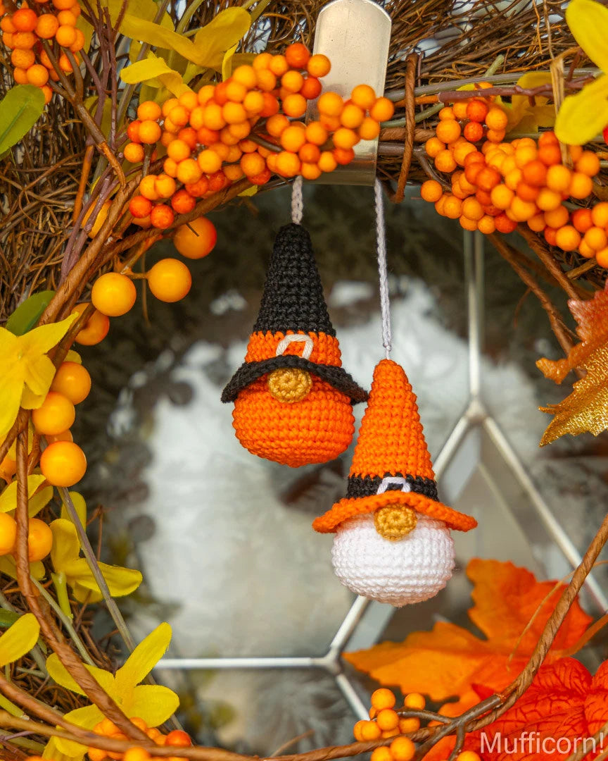 Two crocheted orange and black witch hats hang from a decorative autumn wreath made of twigs, orange berries, and fall leaves. One hat has a black pointed top and the other has an orange pointed top, both with small white and gold details.