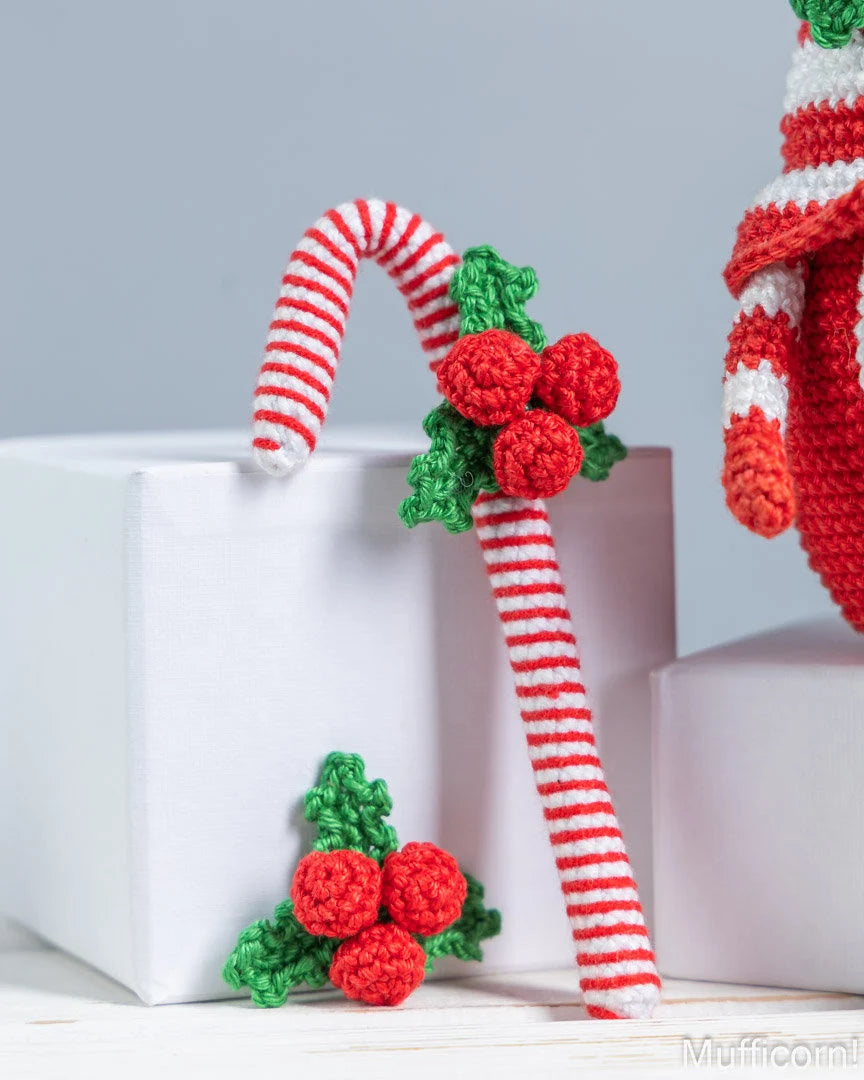 Crocheted Christmas candy cane ornaments with red and white stripes, green holly leaves, and red berries, displayed on white gift boxes against a light gray background.