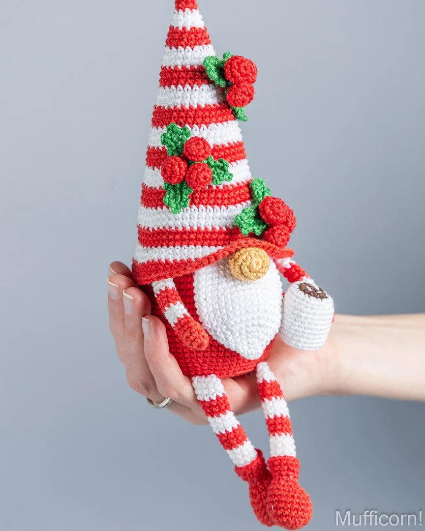A hand holds a crocheted Christmas gnome doll with a red and white striped hat decorated with green holly and red berries, a white beard, and red-and-white striped legs, against a plain gray background.