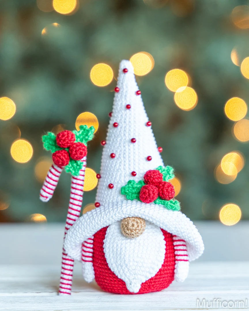 A crocheted Christmas gnome decoration with a tall white pointed hat decorated with small red beads, a red and white striped body, a white beard, and red holly accents, holding a candy cane against a blurred festive background with warm yellow bokeh lights.