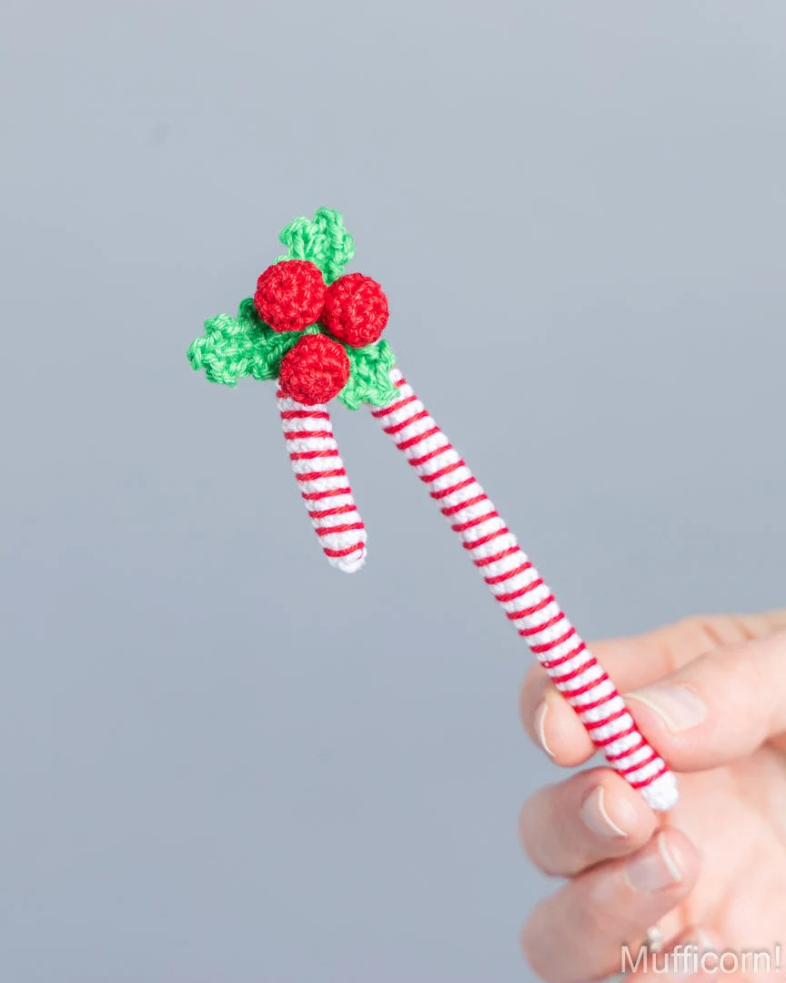 A hand holding a crocheted Christmas-themed candy cane featuring red-and-white striped handles, green holly leaves, and three red berries on a plain gray background.