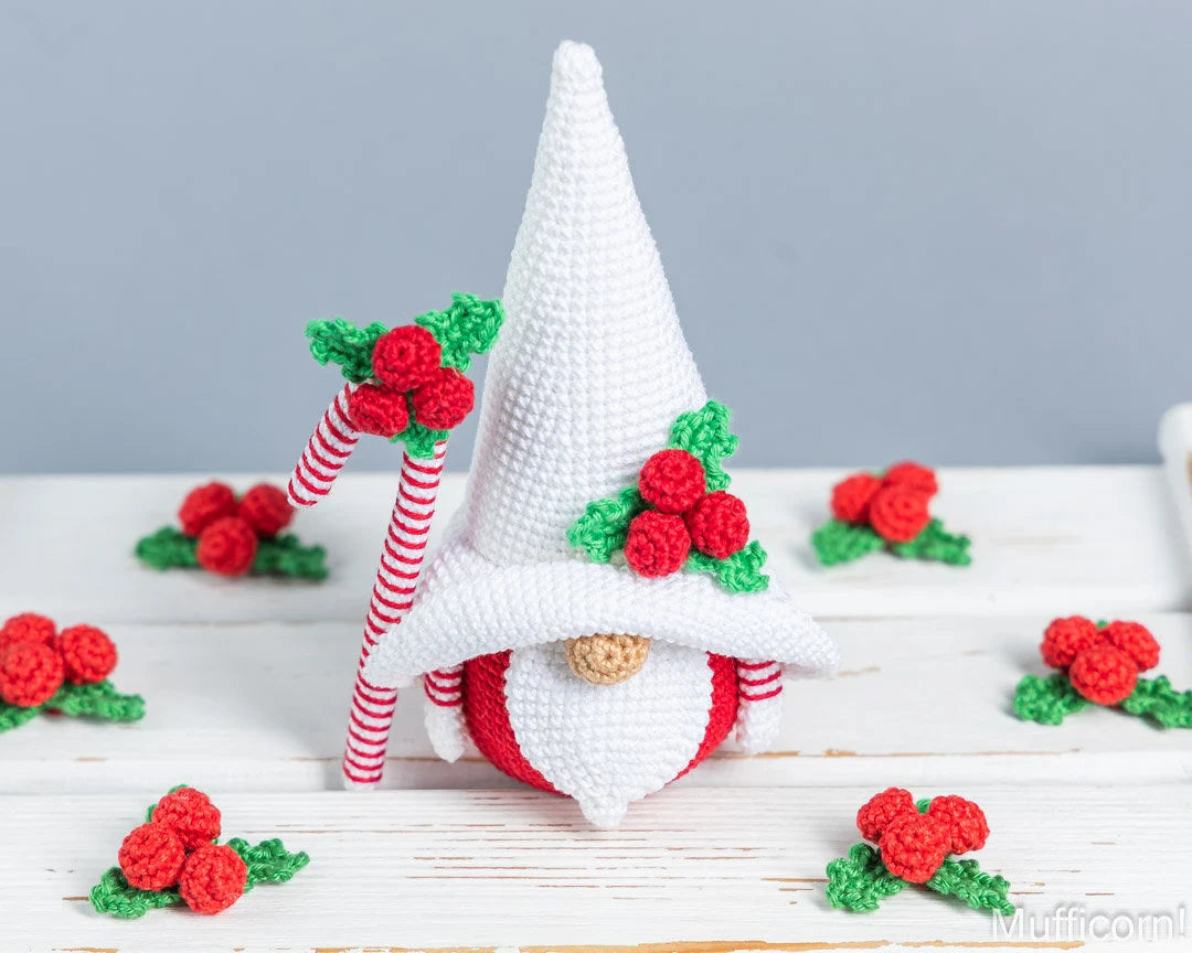 A crocheted Christmas gnome figurine with a tall white pointed hat, red and white striped cane, and red berries with green leaves, displayed on a white wooden surface with smaller matching berry clusters around it.