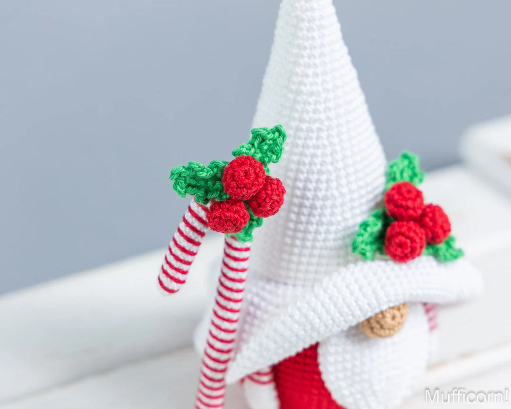 A close-up of a crocheted Christmas gnome figure wearing a tall white pointed hat and holding a red-and-white striped candy cane decorated with green holly leaves and red berries.