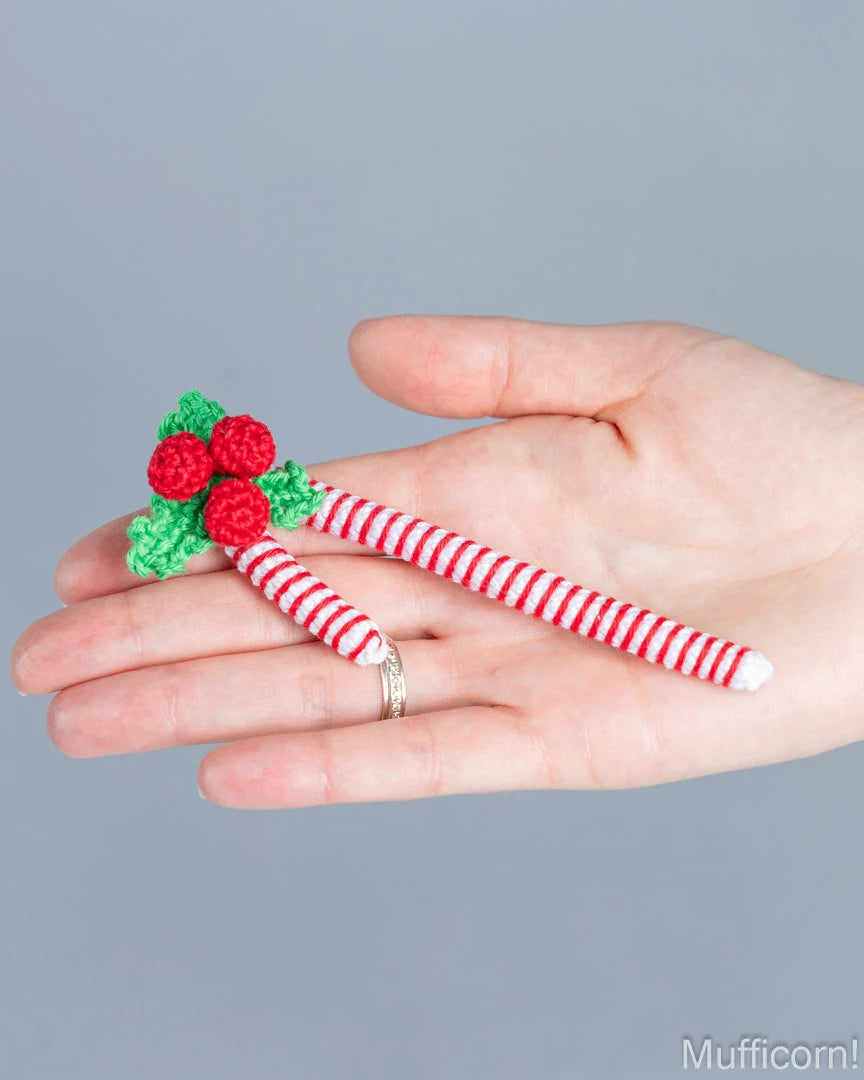 A hand holding two small crocheted candy cane pins with red and white stripes and green holly leaves with red berries, against a solid gray background.