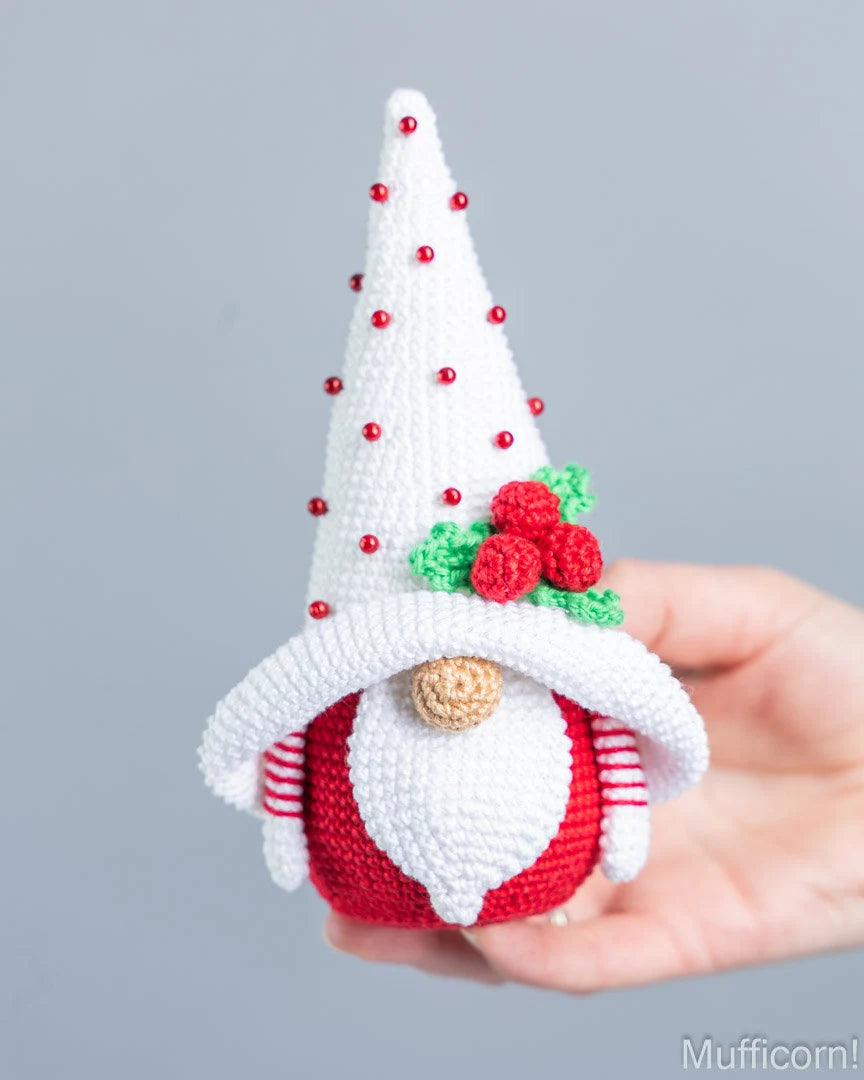 A crocheted Christmas gnome doll being held in a person's hand against a plain light gray background. The gnome has a tall white pointed hat with small red bead accents, a red and white striped body, a white beard, and a green and red holly decoration on the hat.