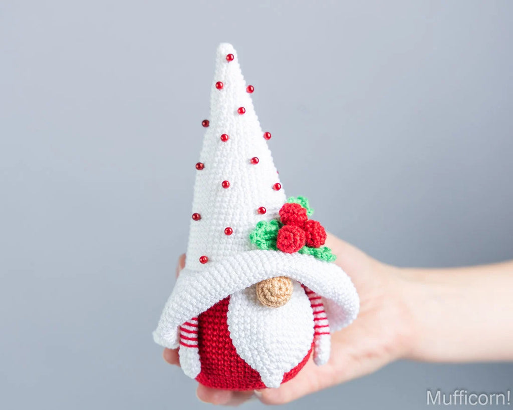 A hand holding a crocheted Christmas gnome doll with a tall white hat decorated with small red beads, a red and white striped body, and a green holly accent.