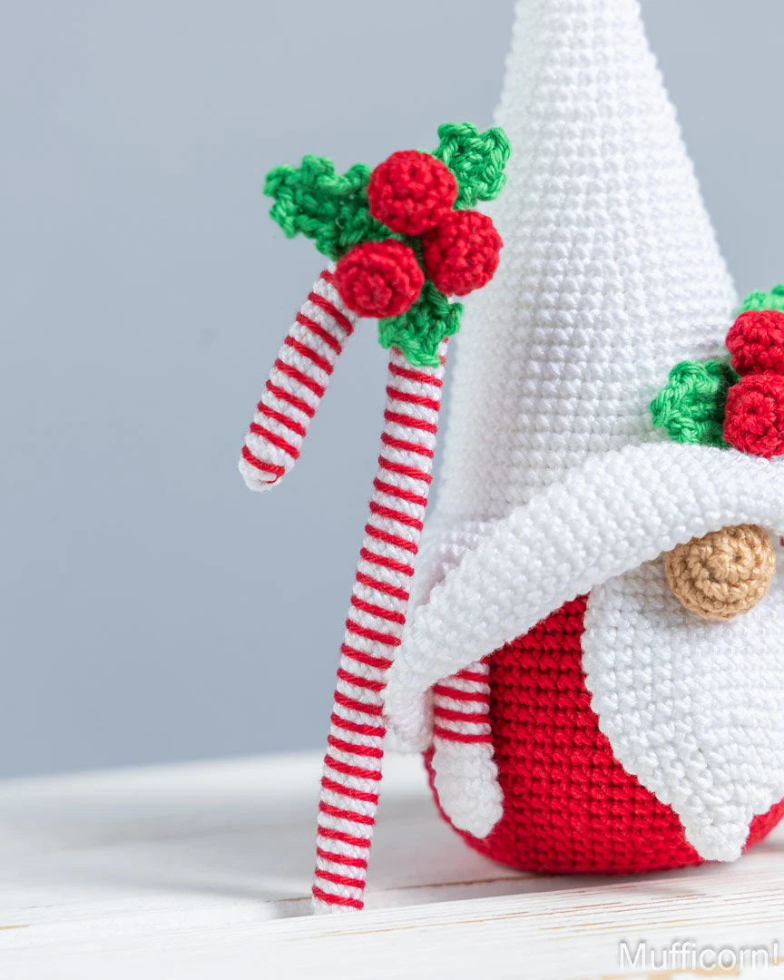 A crocheted Christmas gnome doll with a tall white hat, red and white striped cane, and red and green holly details, photographed on a light surface against a soft gray background.