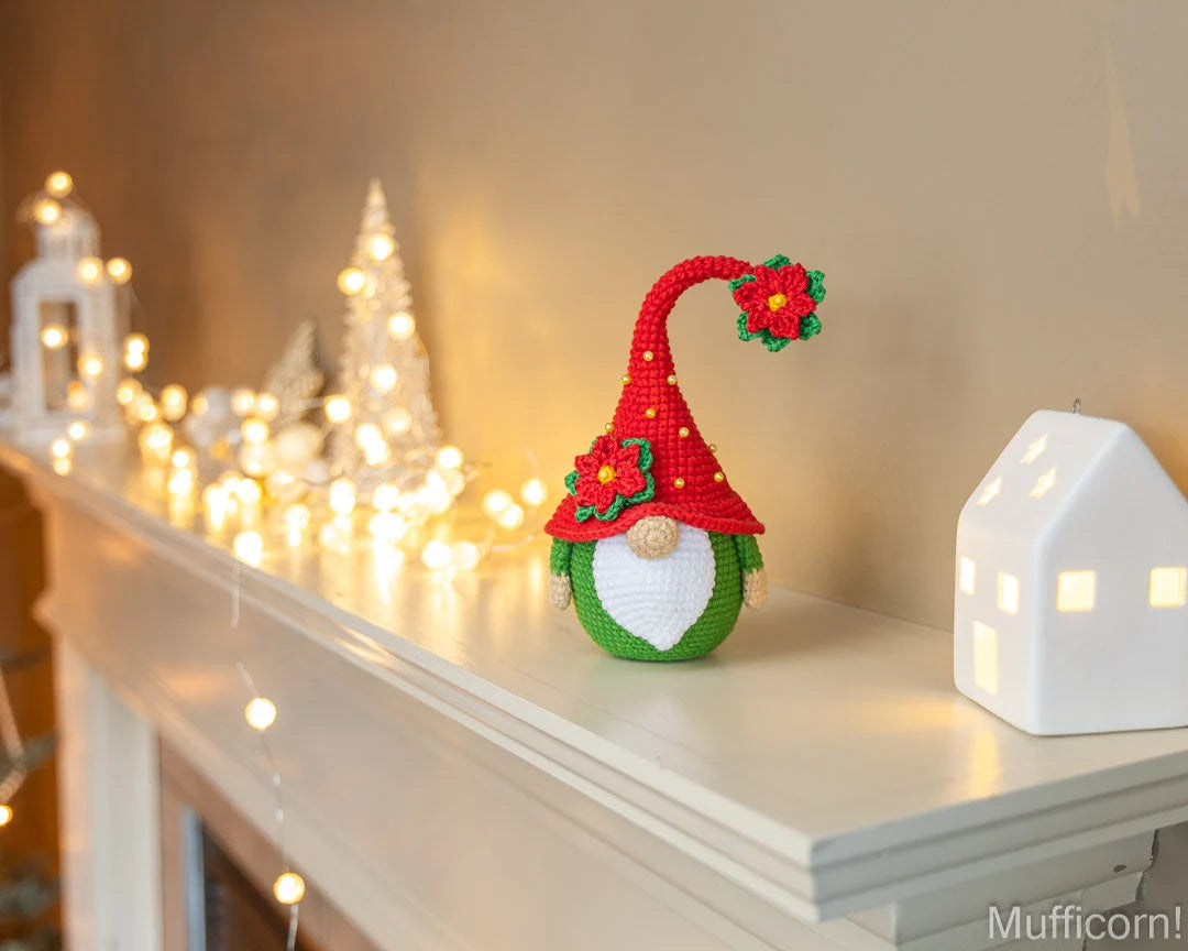 A crocheted Christmas gnome ornament with a red hat, green body, white beard, and red-and-green floral accents, displayed on a white mantel beside warm white string lights and a white house-shaped lamp.
