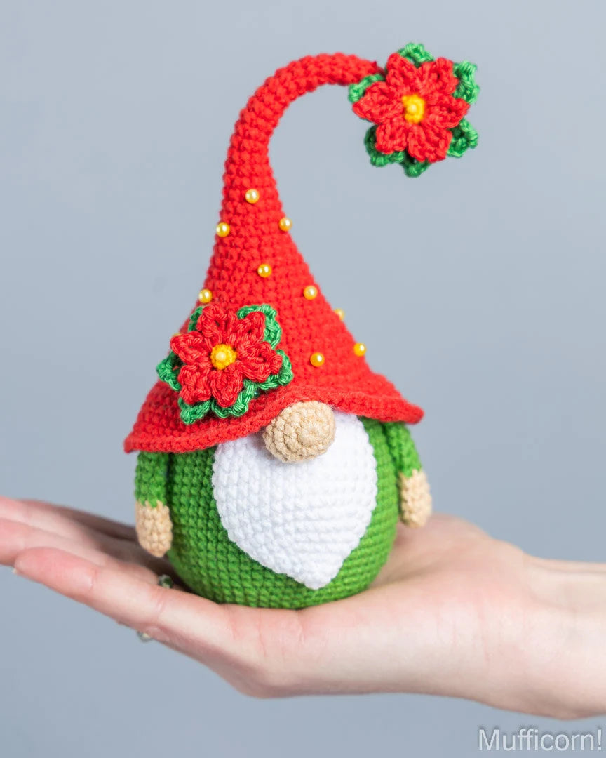 A crocheted Christmas gnome doll held in a person's hand against a plain light gray background. The gnome has a red pointed hat with red poinsettia flowers and gold beads, a green body, a white beard, and a beige nose.