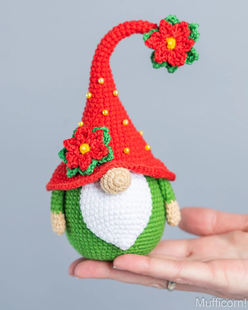 A crocheted Christmas gnome ornament being held in a person's hand against a plain gray background. The gnome has a red pointed hat with red poinsettia flowers, a white beard, green body, and small gold bead accents.