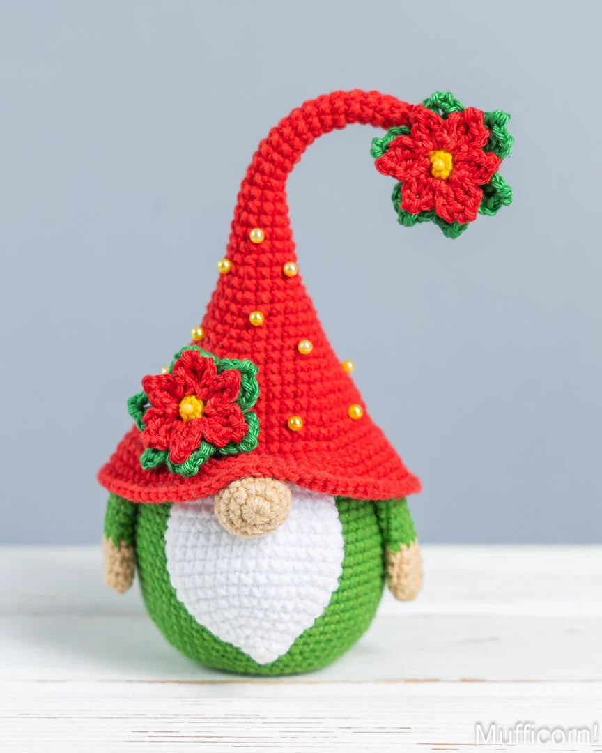 A crocheted Christmas gnome doll with a red pointed hat decorated with red poinsettia flowers and small gold beads, a green body, a white beard, and tan hands and nose, standing on a white surface against a light gray background.
