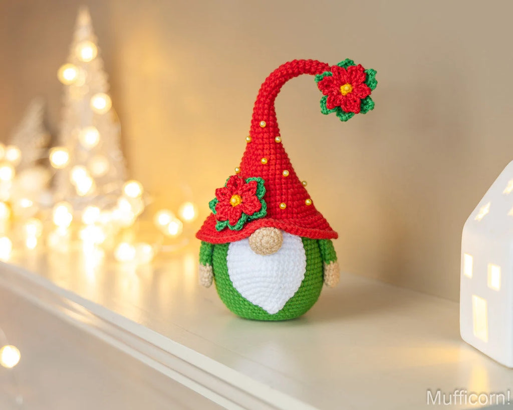 A crocheted Christmas gnome figurine with a red pointed hat, green body, white beard, and red flower accents, displayed on a shelf in a warmly lit indoor setting.
