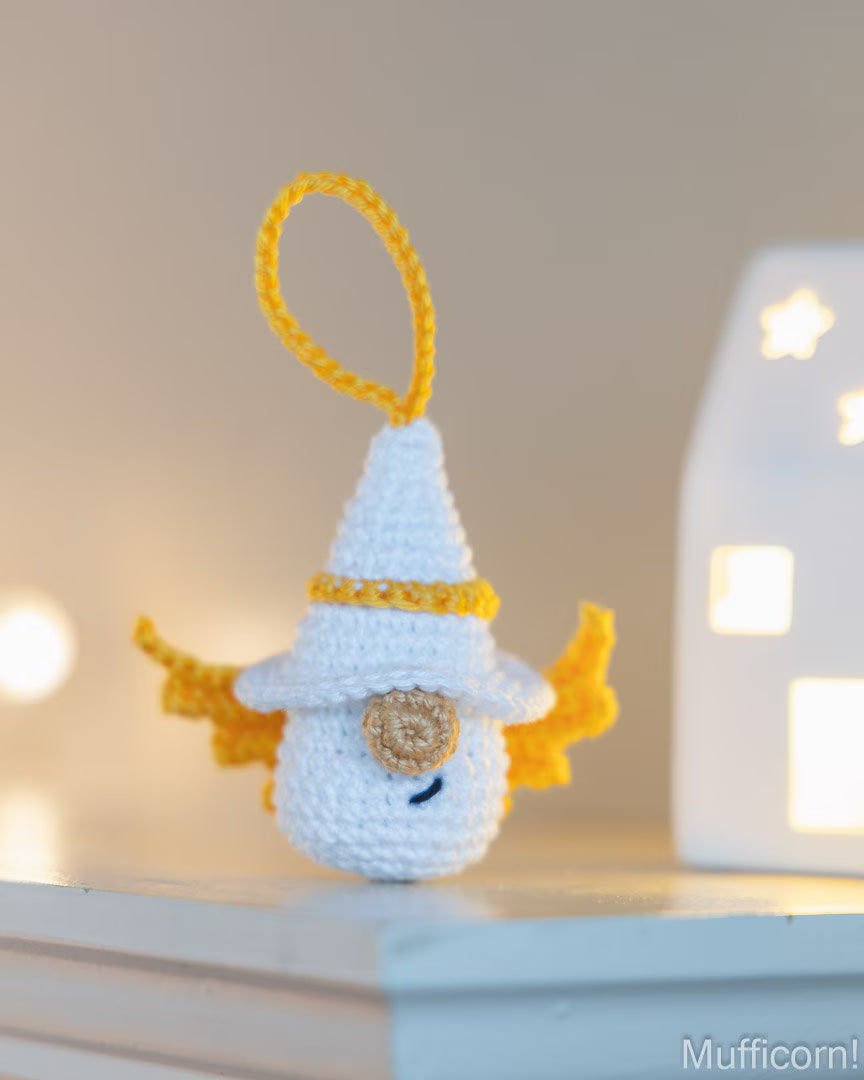 A small crocheted white angel ornament with yellow wings, a yellow loop at the top, and a gold button on the chest, displayed on a shelf with a softly blurred warm background and a glowing white house light.