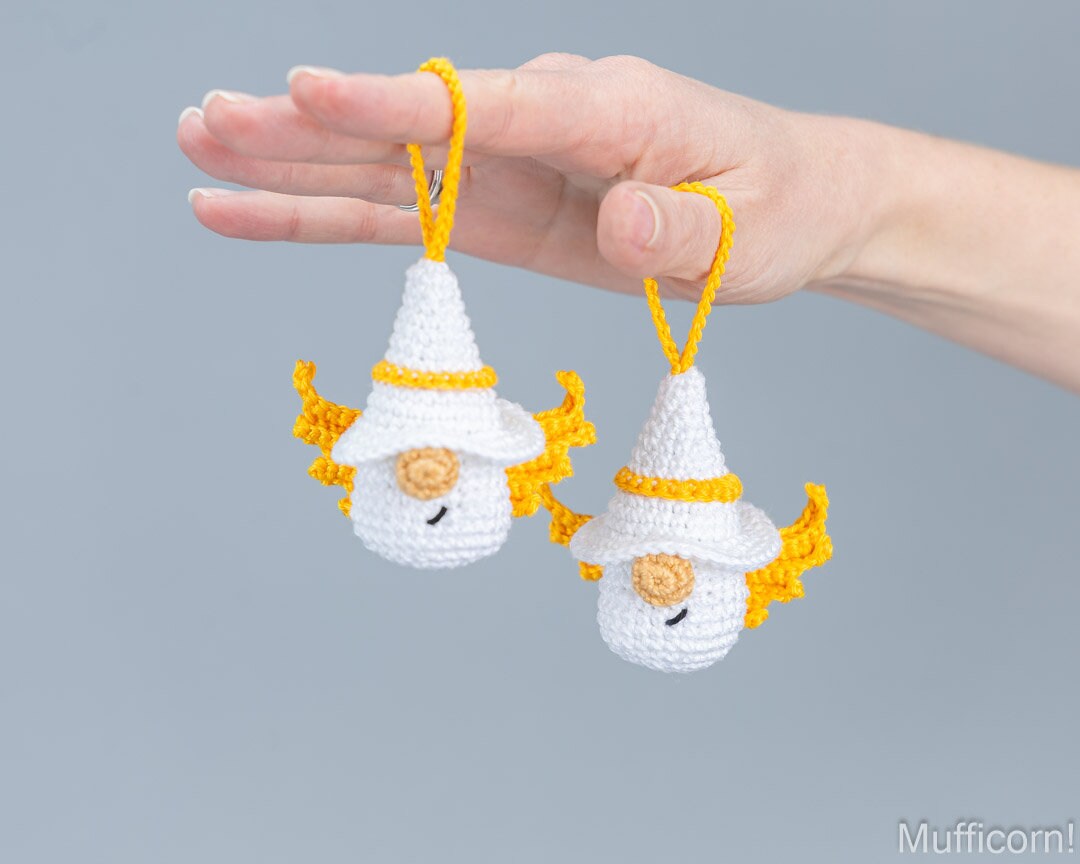 Two small crocheted gnome angels ornaments are held in a person's hand against a plain gray background. The ornaments are white with yellow loops and yellow accents, and have embroidered smiles.