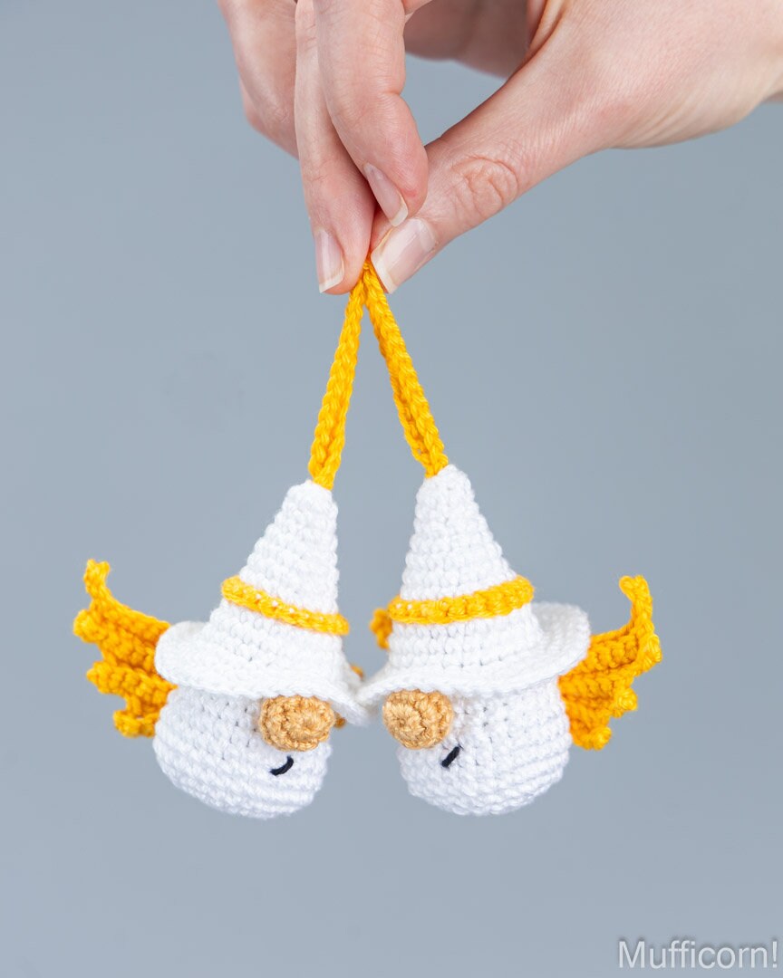 A pair of white crochetedangel gnomes being held by a person's hand, suspended by yellow strings against a plain gray background.
