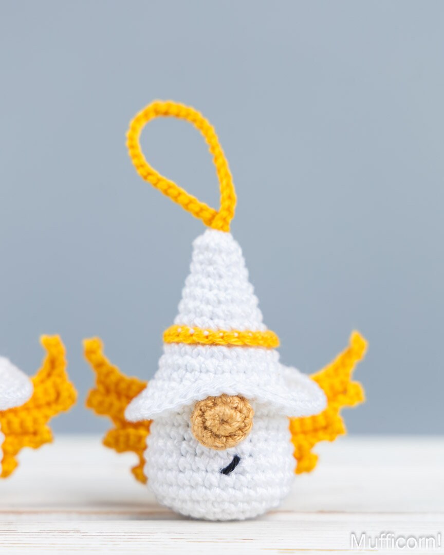 A small crocheted white and yellow angel gnome ornament with a pointed hat, yellow wings, and a yellow hanging loop, shown on a light surface against a soft gray background.