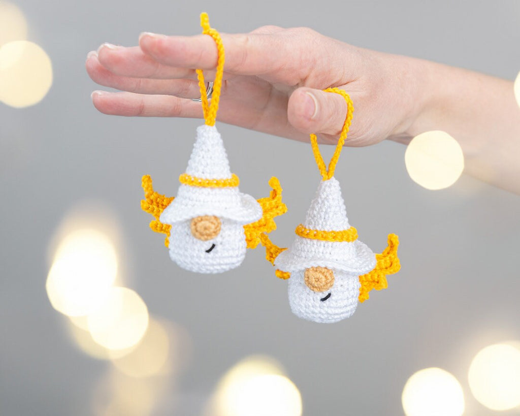 A hand holds two small crocheted gnome ornaments with white hats, yellow trim, and yellow wings against a soft gray background with warm bokeh lights.
