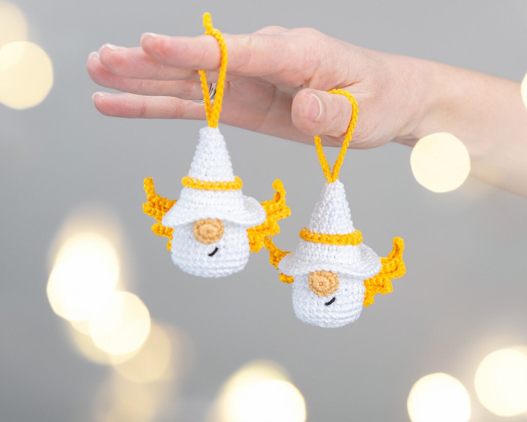 A hand holds two small crocheted gnome ornaments with white hats, yellow trim, and yellow wings against a soft gray background with warm bokeh lights.