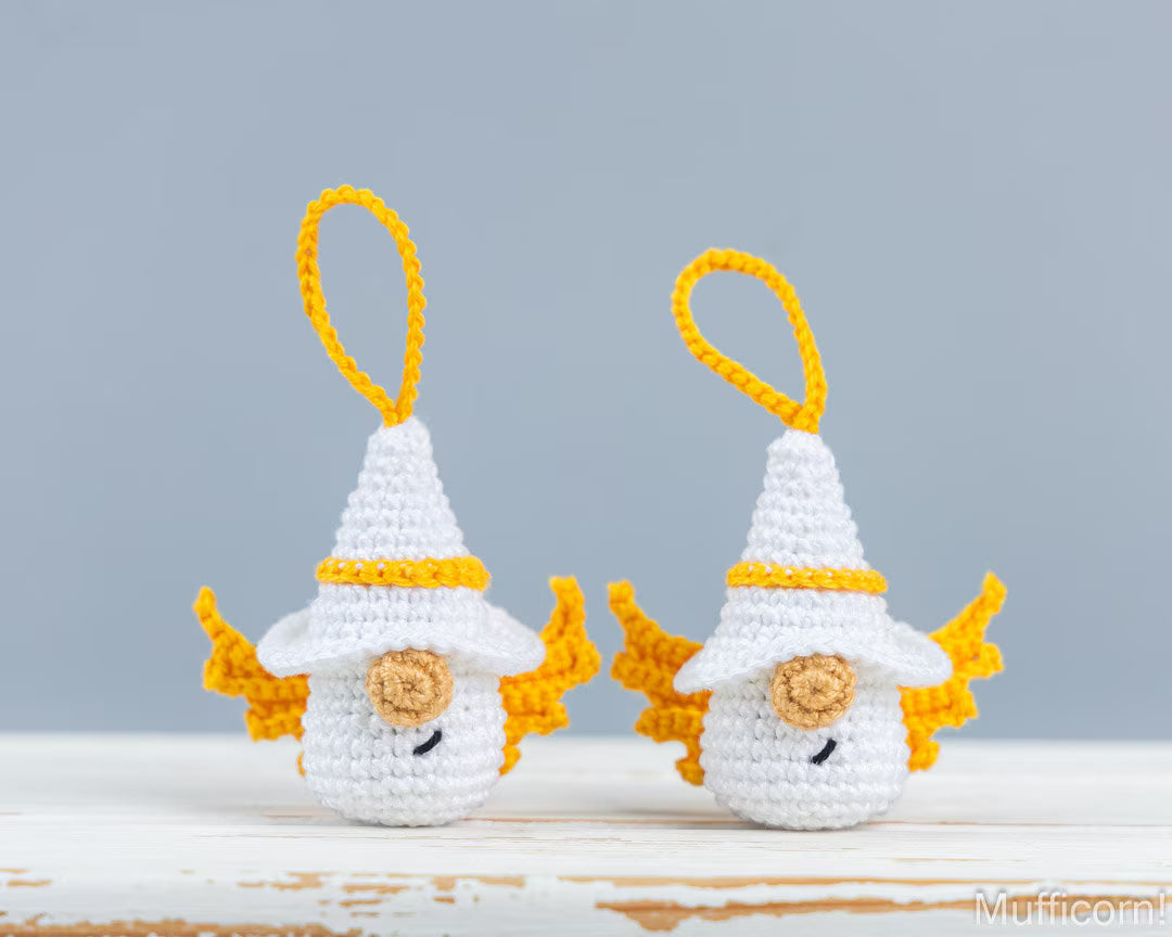 Two small crocheted fairy ornaments with white bodies, yellow wings, and yellow hat loops, displayed on a light surface against a soft gray background.