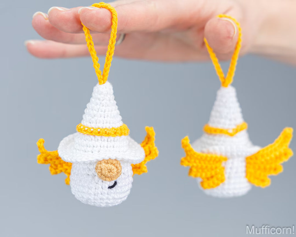 A hand holds two small crocheted hanging angel ornaments, each white with yellow loops and yellow wing-like accents, against a soft gray background.